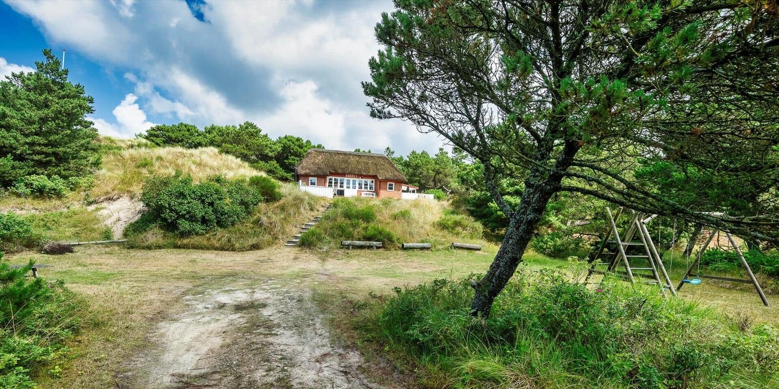 Haus mit Dachrinne auf Dünenhügel, umgeben von Bäumen und Gras.