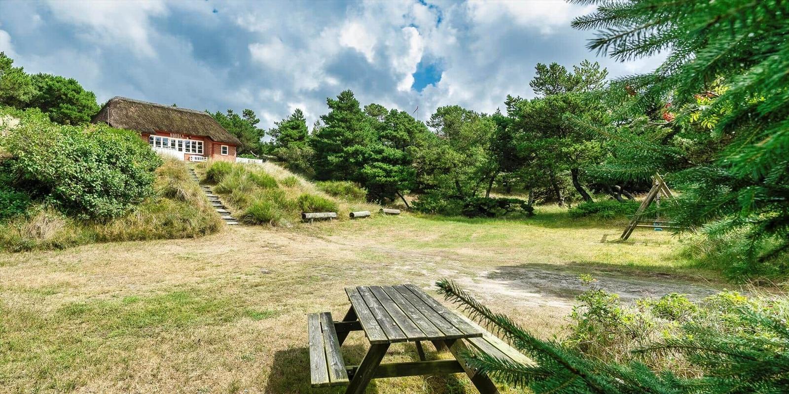 Holzpicknicktisch vor einem roten Haus mit Strohdach. Grünflächen und Bäume im Hintergrund.