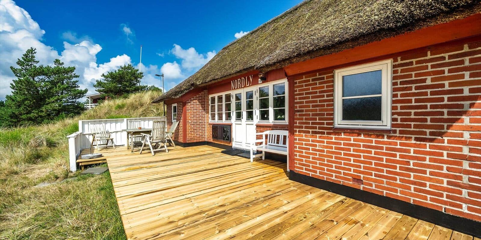 Rotes Backsteingebäude mit strohgedecktem Dach und Holzterrasse mit Sitzgelegenheiten.