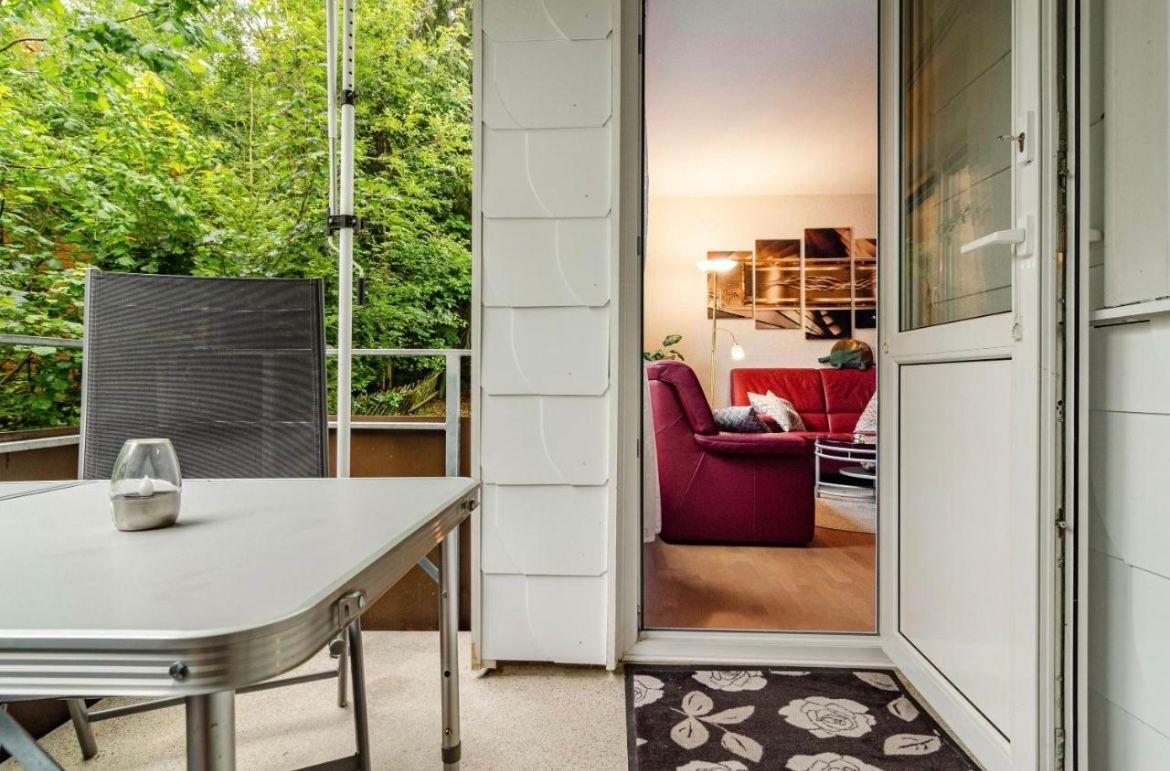 Terrasse mit Tisch und Stuhl. Durch Glastür ist Wohnzimmer mit rotem Sofa sichtbar.