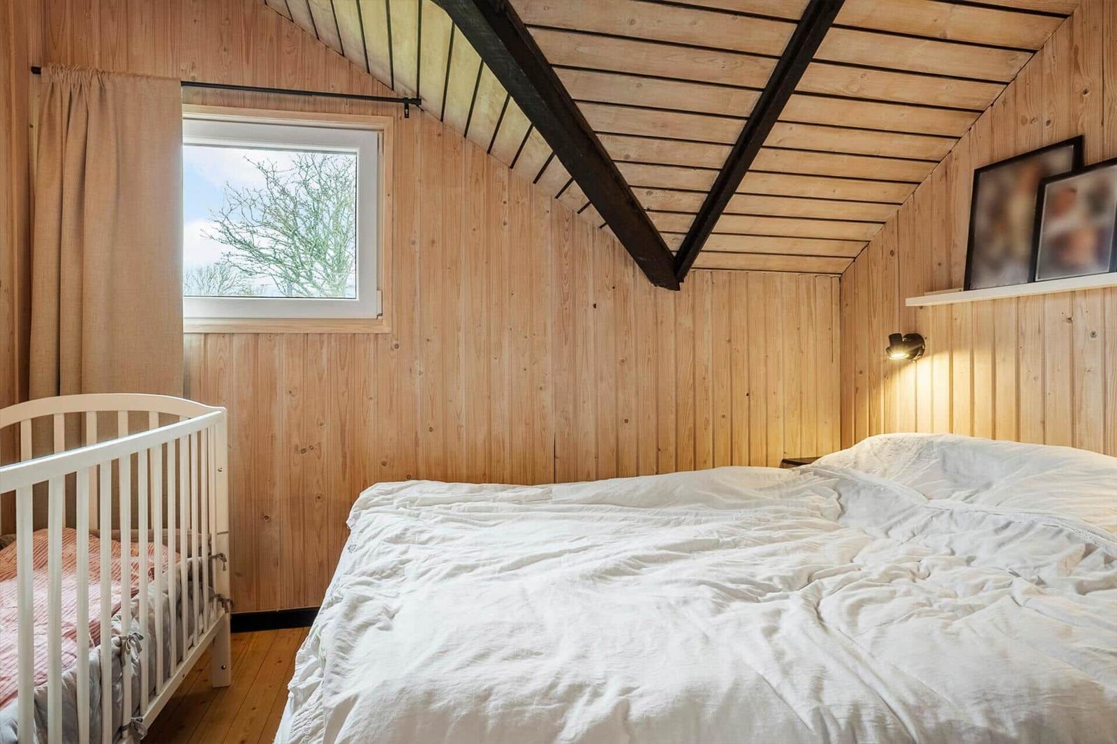 Schlafzimmer mit Holzwand, Bett und Kinderbett. Fenster mit Blick auf Bäume.