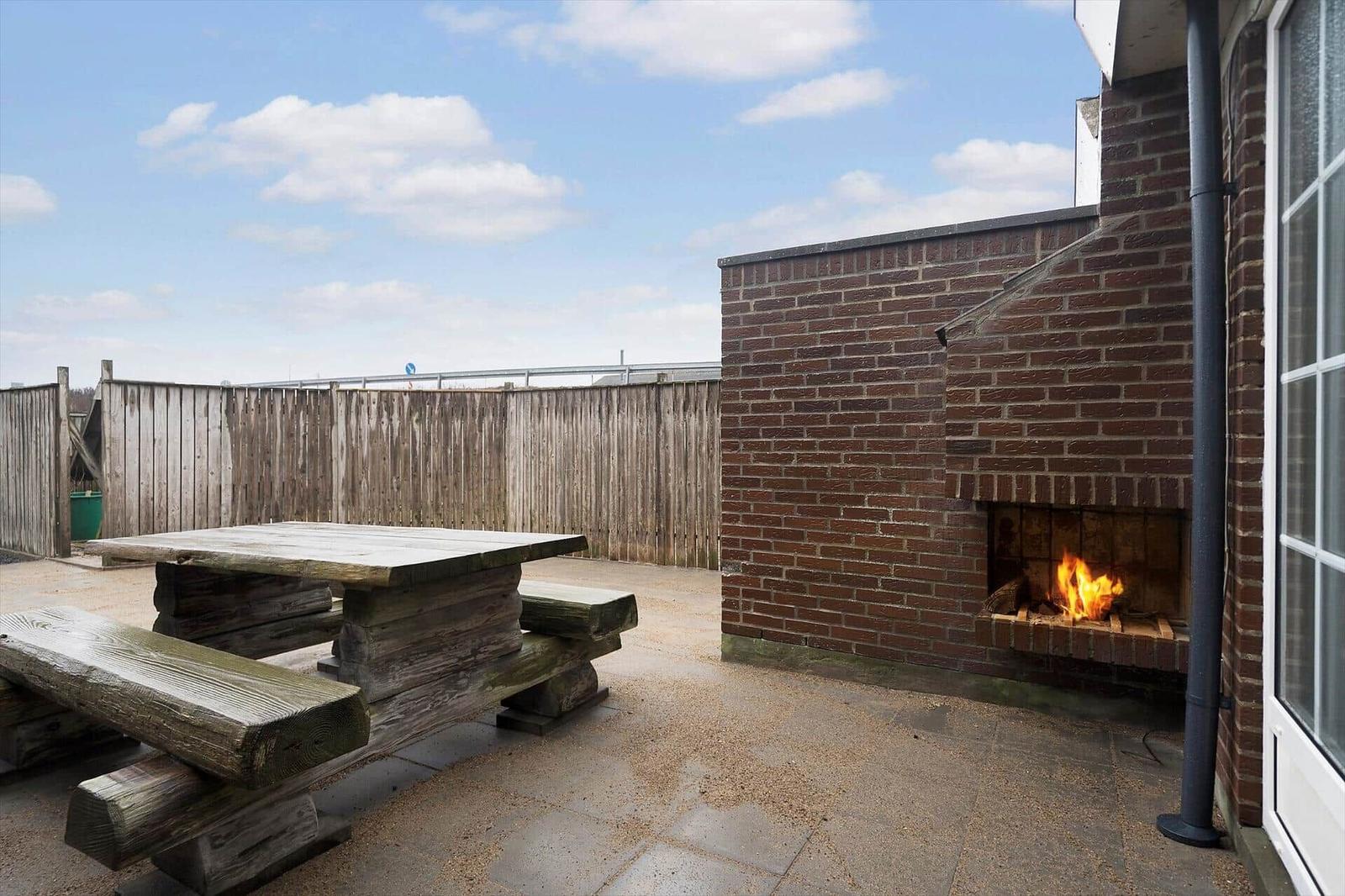 Außenbereich mit Holztisch, Grill und Steinplatten. Hintergrund: Holzzaun und blauer Himmel.