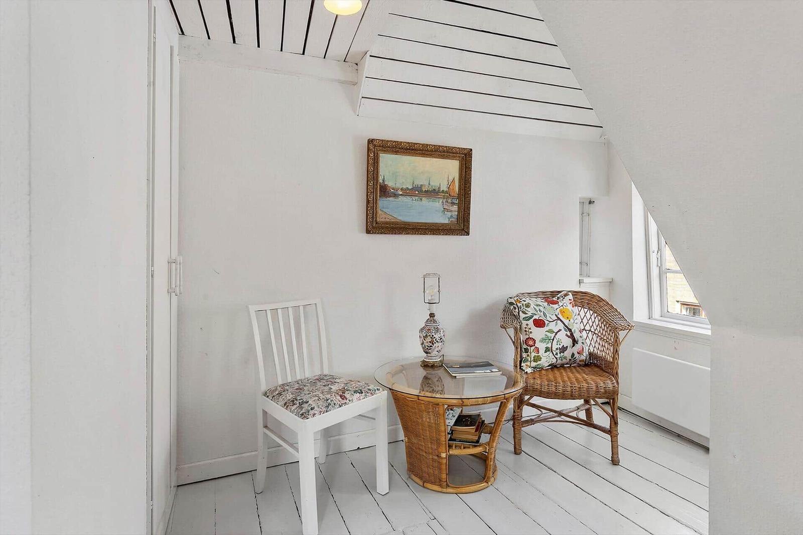 Room with white walls, wooden floor, and sloped ceiling beams. A round table and two chairs are in the room.