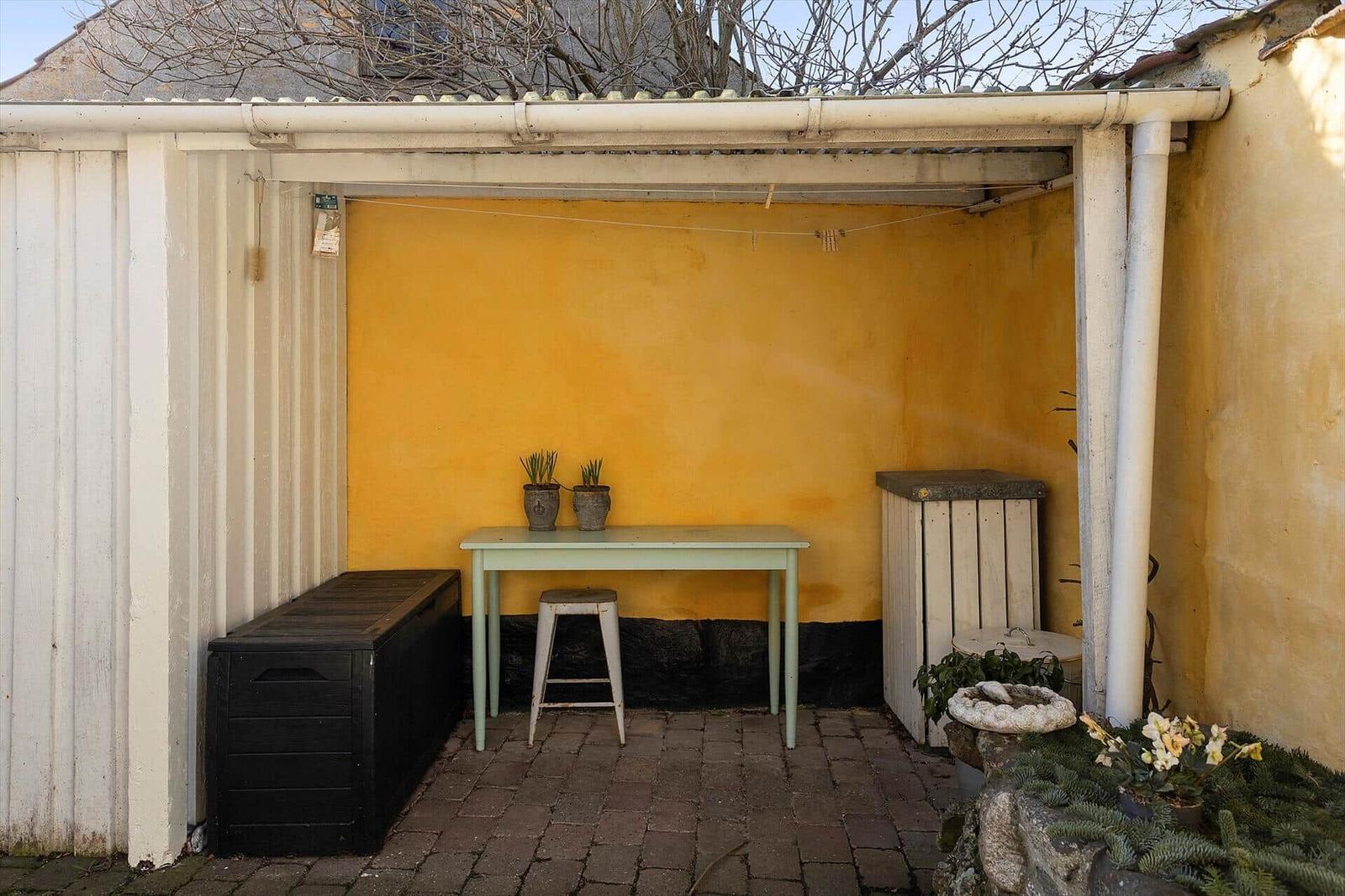 Yellow outdoor area with table, stool, and plants under roof.