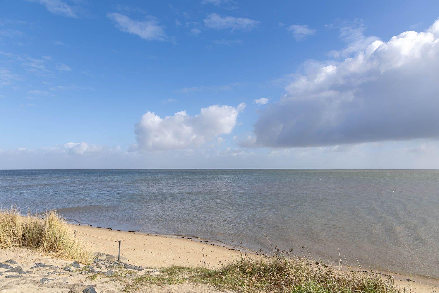 Strand mit Sand, Gras und Meer unter blauem Himmel mit weißen Wolken.