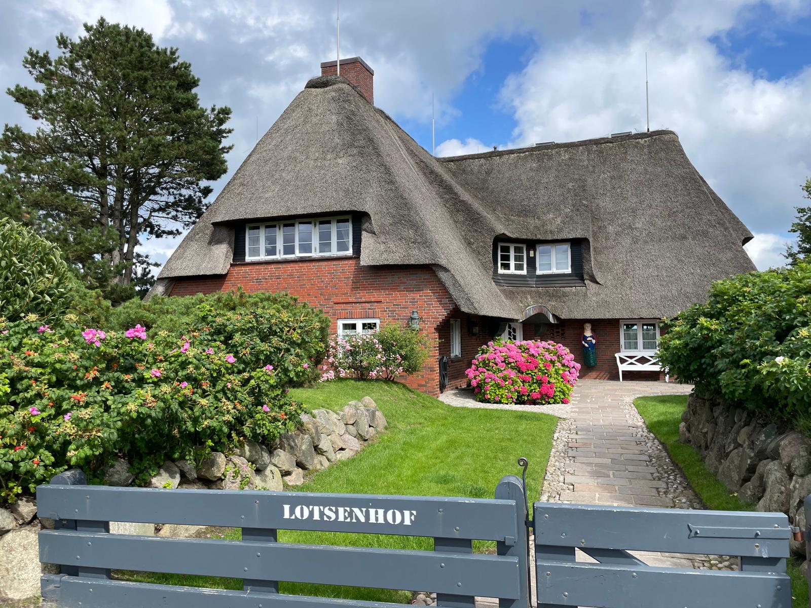 Thatched house with garden path and blooming flowers. Gate reads Lotsenhof.