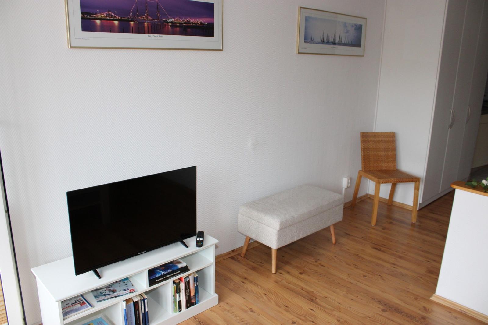 TV auf weißem Regal, Sitzbank und Stuhl im Wohnbereich mit Holzboden und Wandbildern.