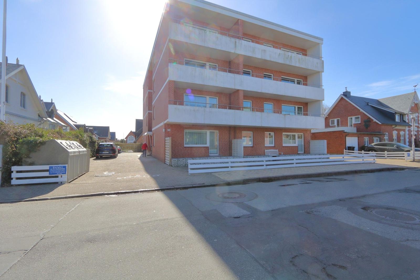 Viergeschossiges Wohnhaus mit Balkonen und weißen Geländern. Straßenansicht mit gepflastertem Weg.