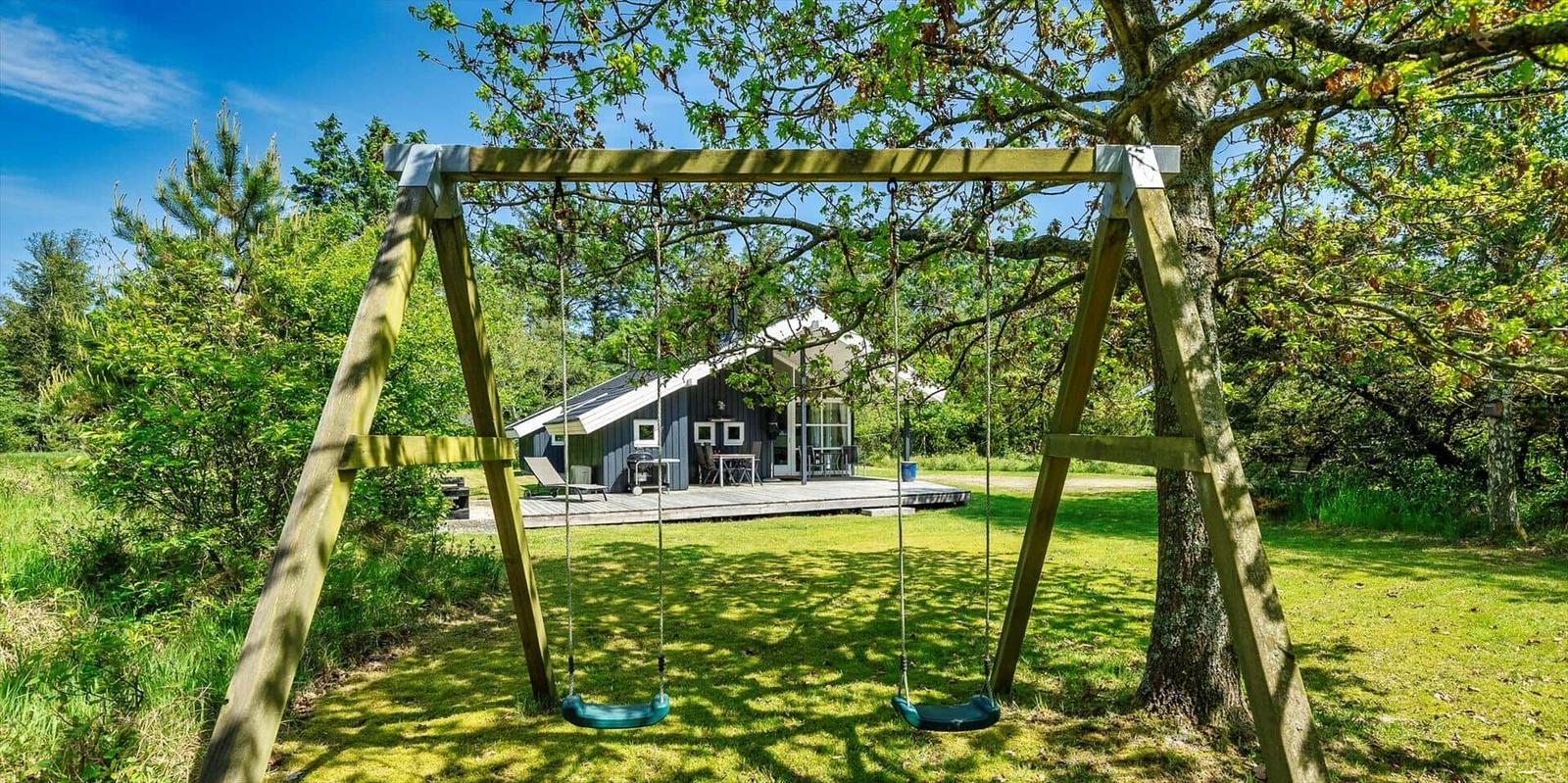 Zwei Schaukeln unter Bäumen vor einem Ferienhaus mit Terrasse.