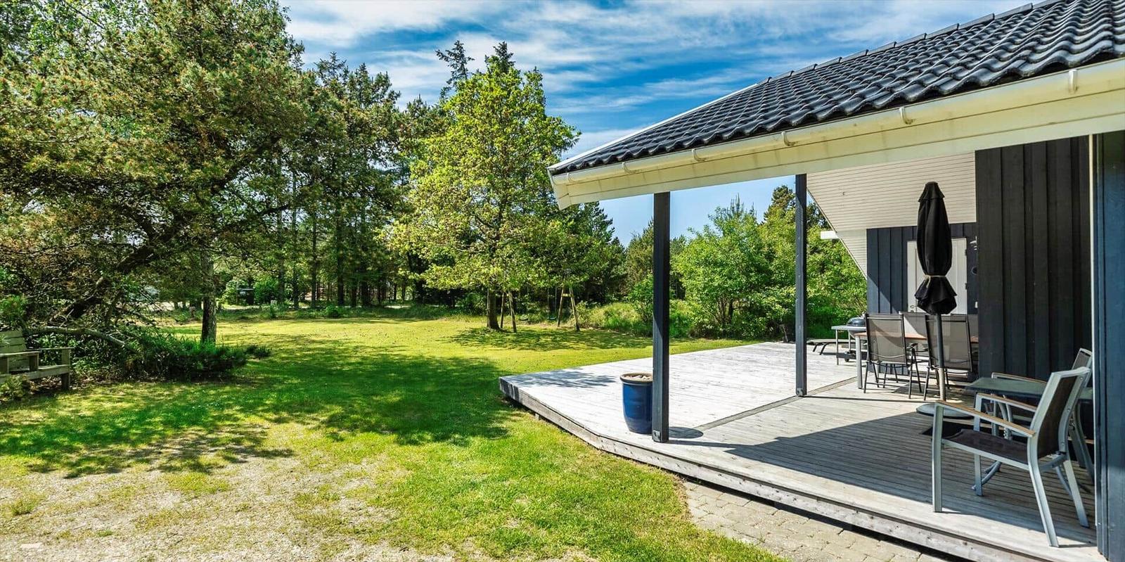Haus mit Terrasse, Garten und Bäumen unter blauem Himmel.