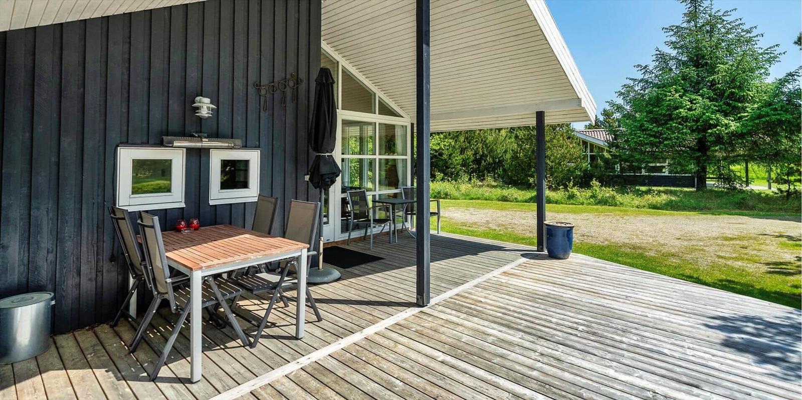 Terrasse mit Tisch, Stühlen und Sonnenschutz. Holzdecke und dunkler Holzwand. Grün hinter dem Haus.
