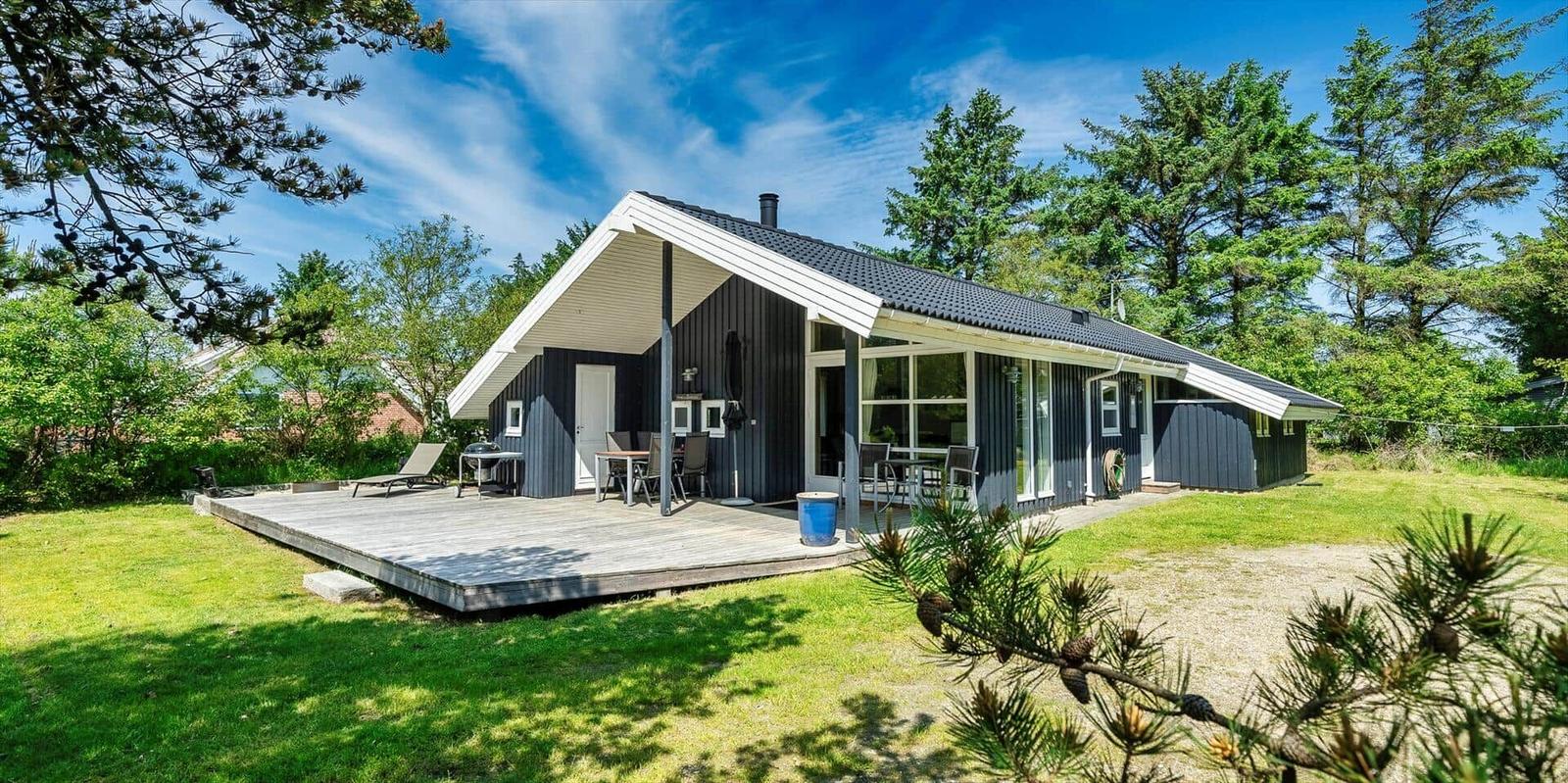 Moderne Holzhütte mit Terrasse, Grill und Garten. Umgeben von Bäumen und Gras.