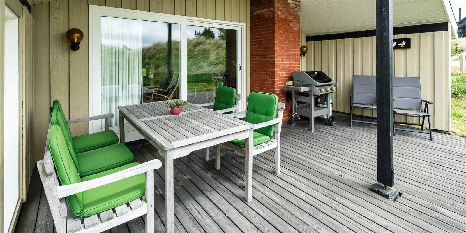 Terrasse mit grünen Sitzkissen, Tisch und Grill. Holzdecke und Glasfront.