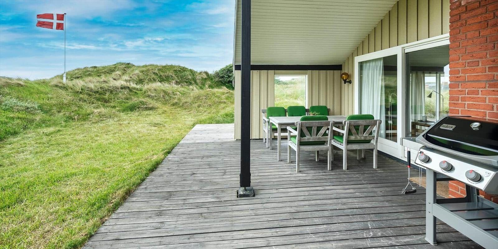 Terrasse mit Grill, Tisch und Stühlen. Grasige Dünen und dänische Flagge im Hintergrund.