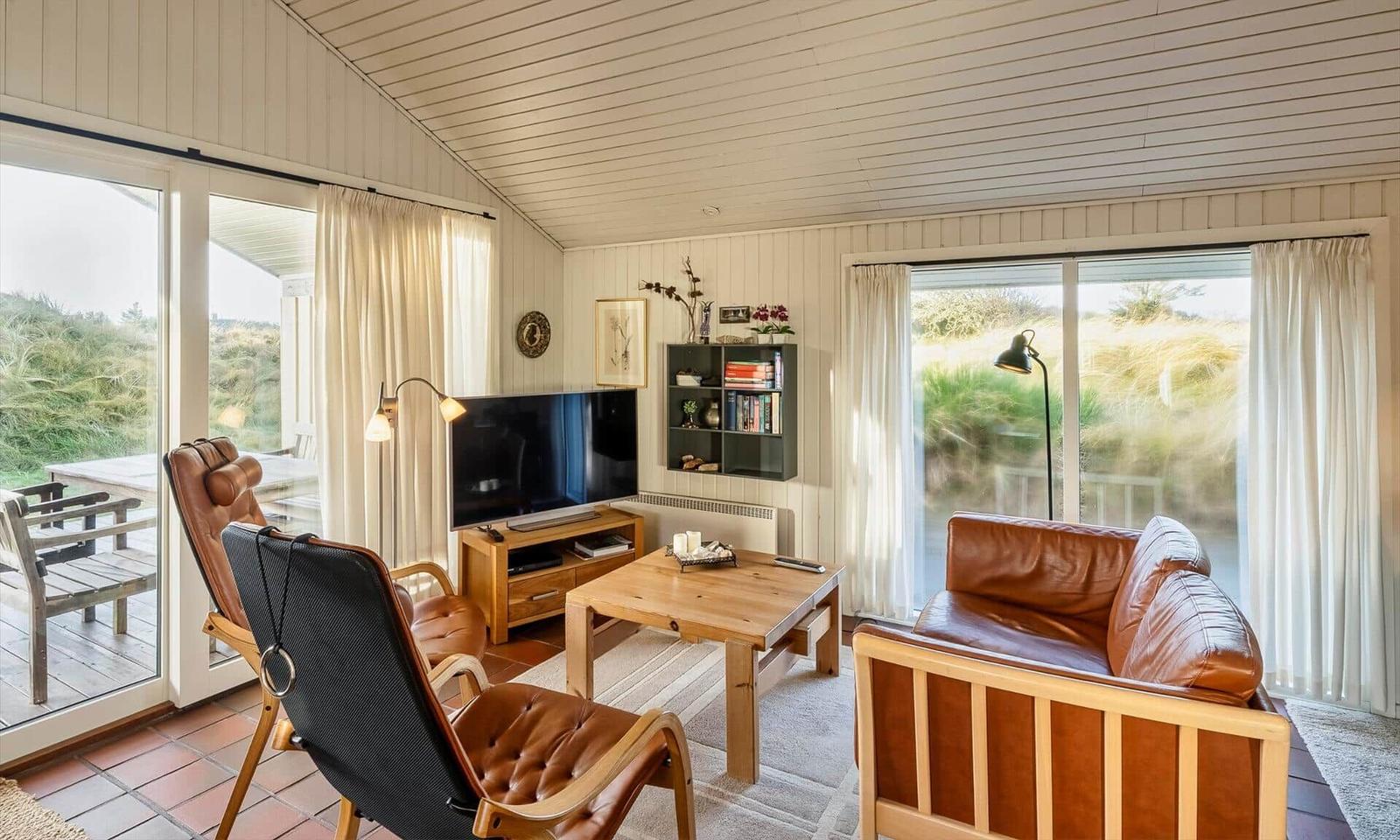 Wohnzimmer mit braunen Ledermöbeln, Holztisch und TV. Große Fenster mit Blick auf Grasland.