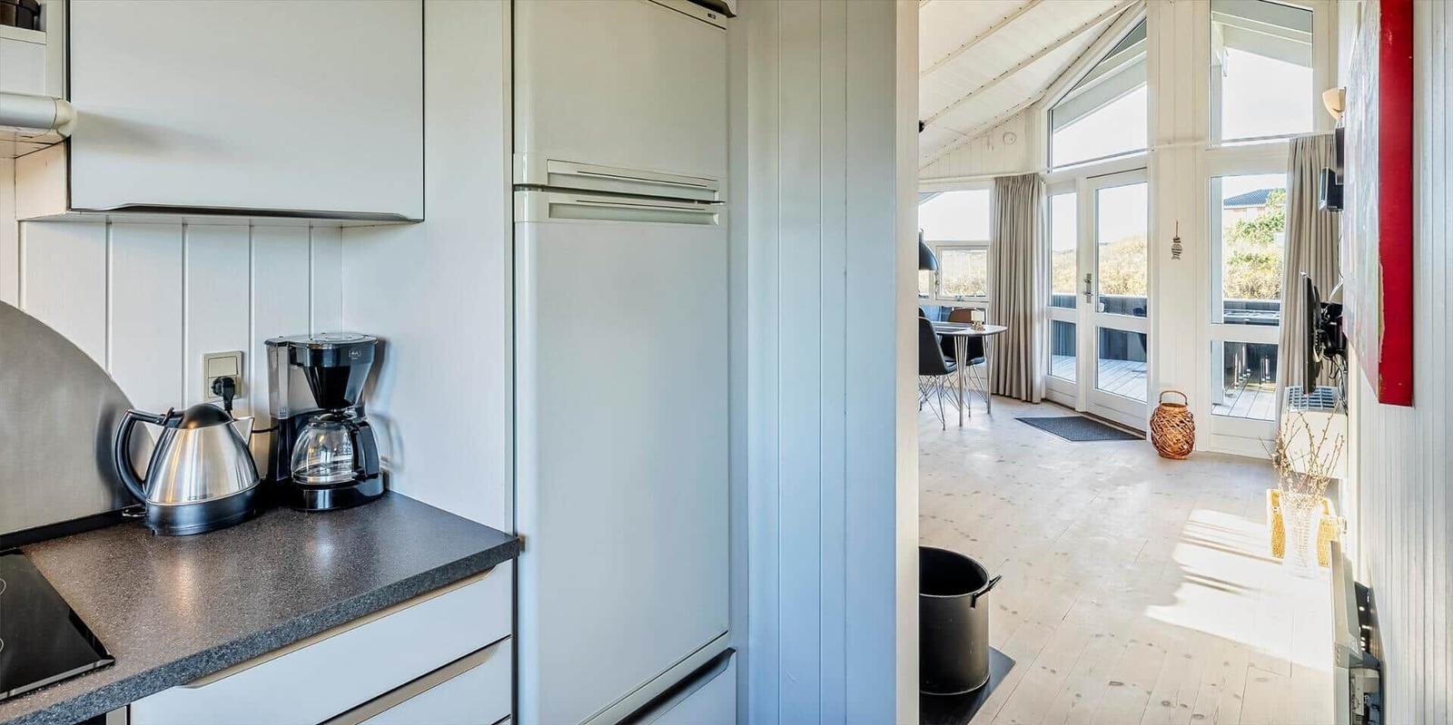 Küche mit Kaffeemaschine und Kühlschrank. Blick in Wohnbereich mit Tisch und Balkontür.