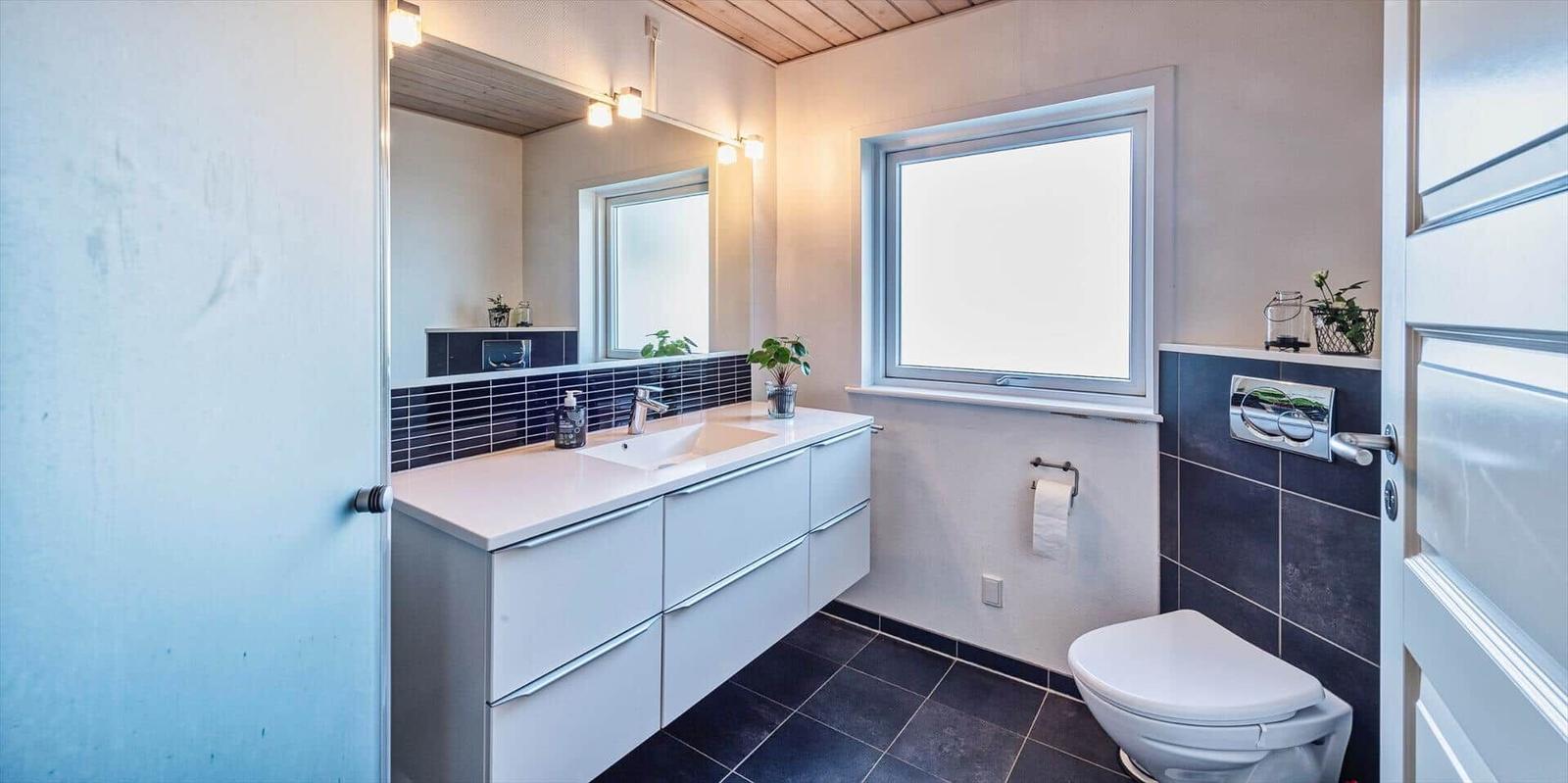 Badezimmer mit Waschbecken, Spiegel und Fenster. Toilette und Fliesenboden.