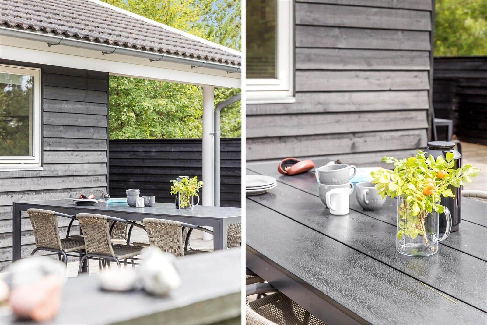 Terrace with table, chairs, plants, and mugs. Gray wooden walls and green trees in the background.