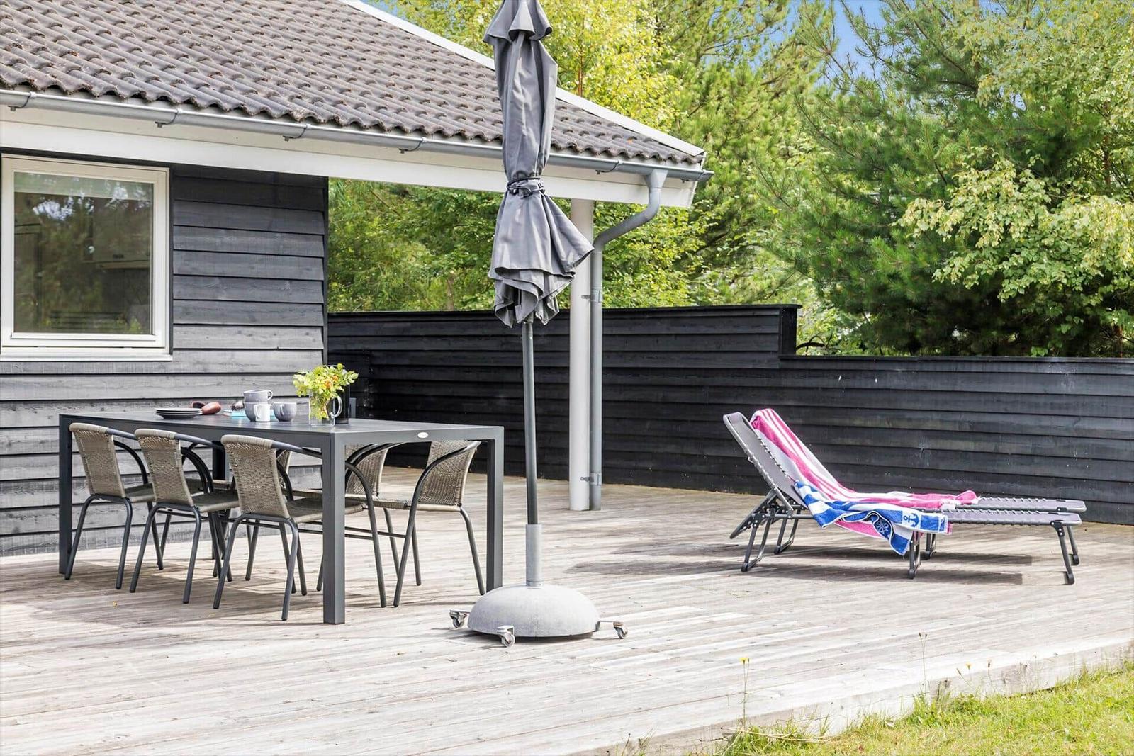 Terrace with table, chairs, and lounge chairs. Background: wooden fence and trees.