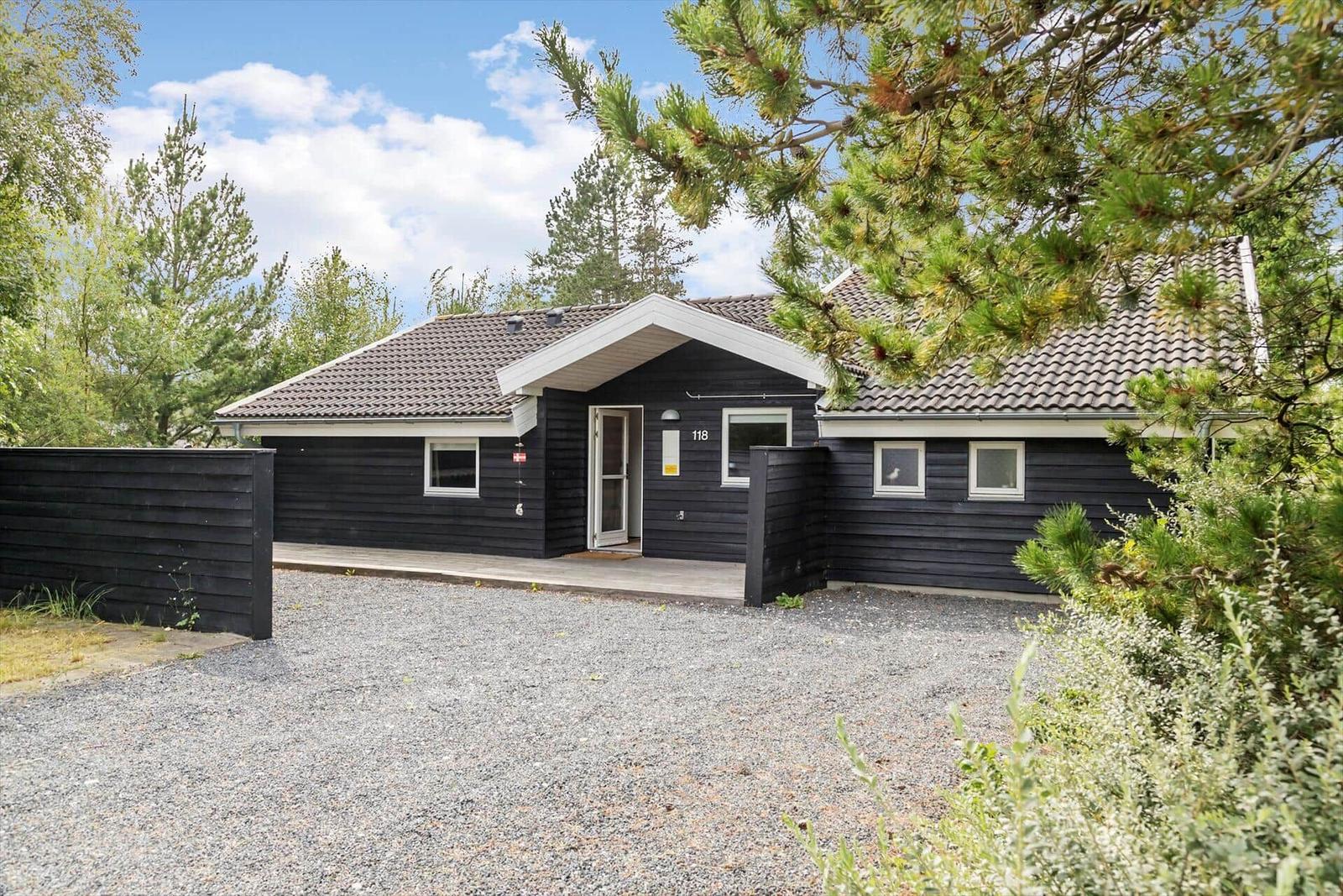 Black wooden house with roof and windows. Front yard with gravel and trees.