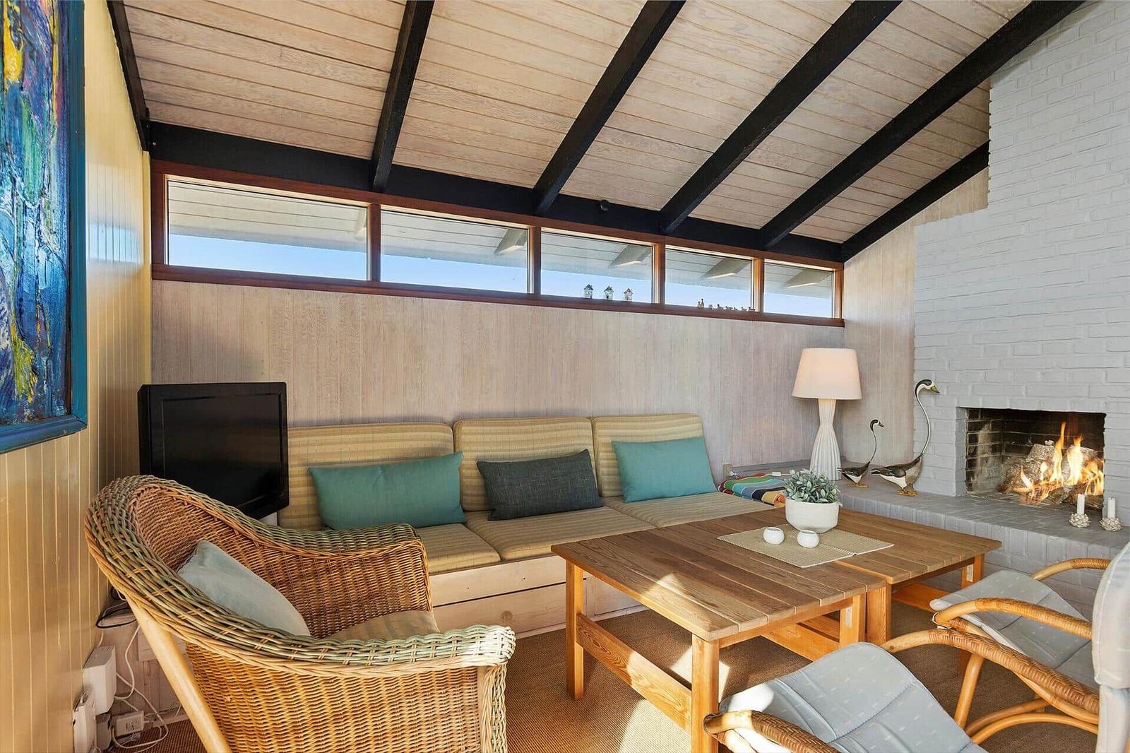 Living room with sofa, fireplace, and wooden table. Ceiling with wood paneling and window row.