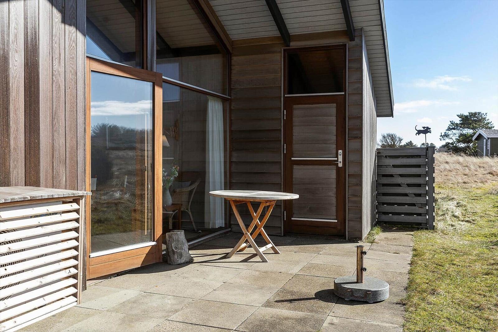 Terrace with table and chair in front of wooden house with glass facade and entrance door.
