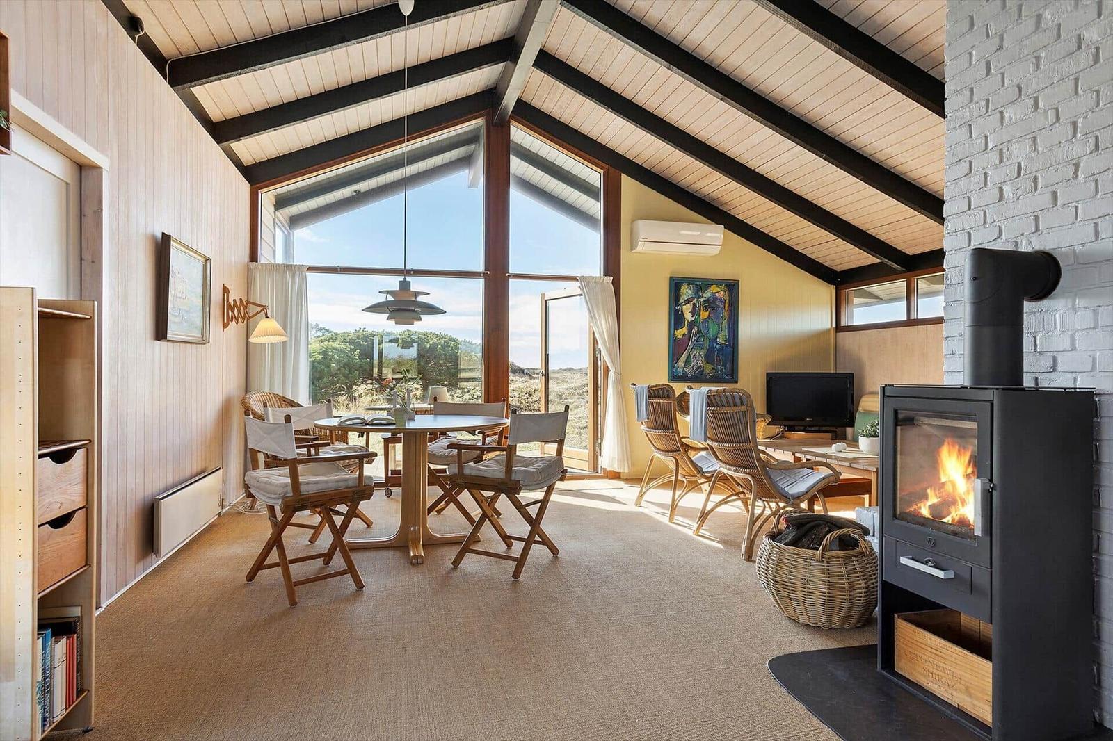 Living and dining area with wooden table, chairs, and wood stove. Large windows with view of landscape.
