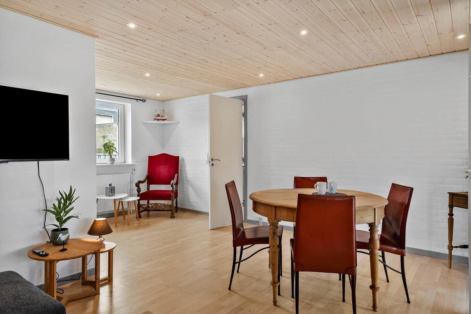 Esszimmer mit runden Tisch und roten Stühlen, daneben Wohnbereich mit Fernseher und roter Sessel.