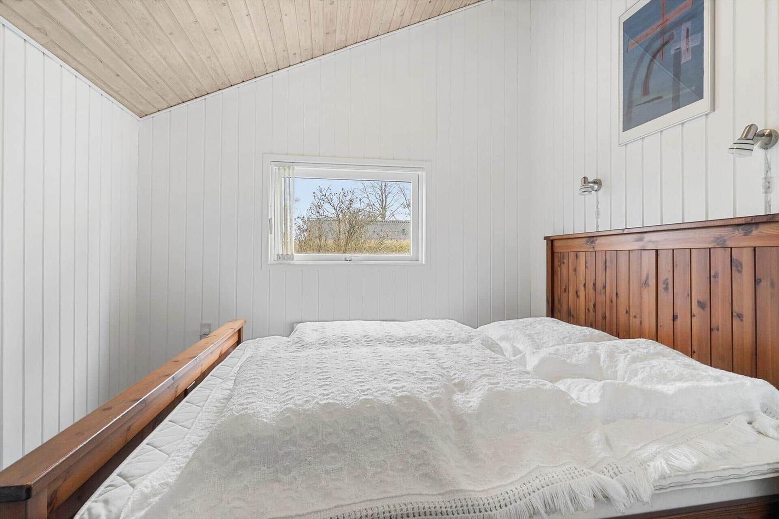 Schlafzimmer mit Holzbett, weißer Wandverkleidung und kleinem Fenster.