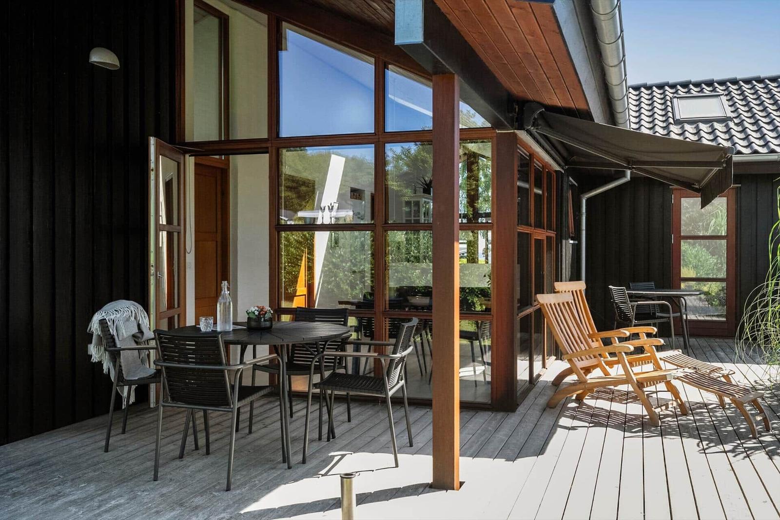 Terrasse mit Tisch, Stühlen und Liegen. Holzdecke und große Fenster.