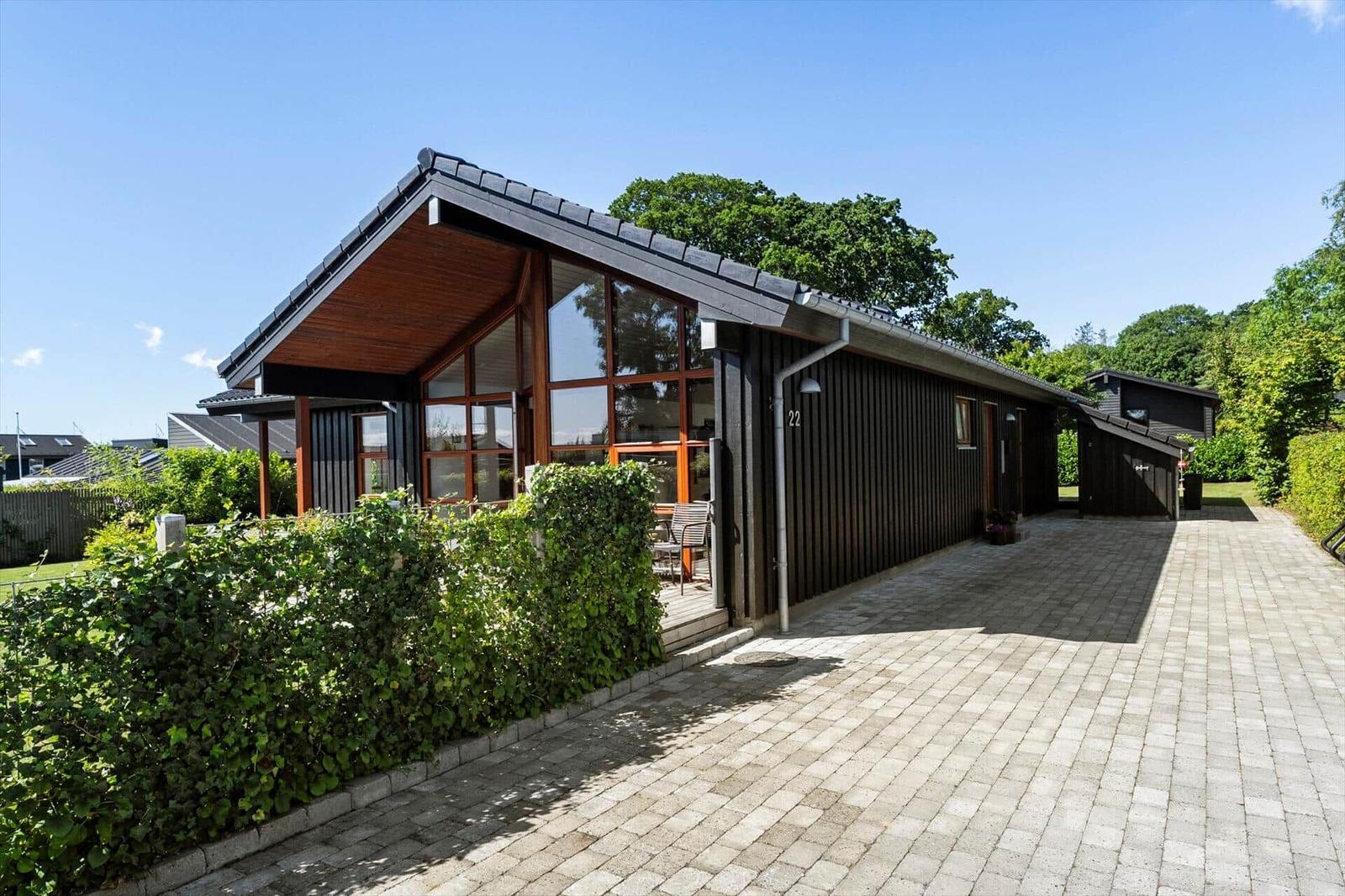 Moderne Holzhütte mit großem Fenster und gepflastertem Hof. Umgeben von grüner Hecke und Bäumen.