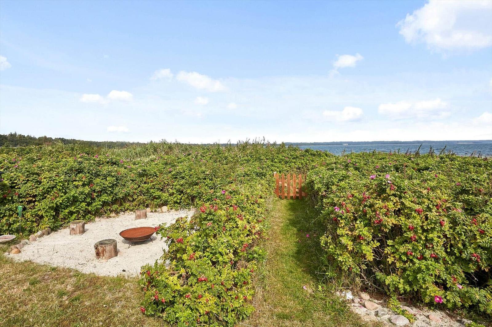 Gartenbereich mit Feuerkorb und Stümpfen neben Blumenhecken und Blick auf Wasser.