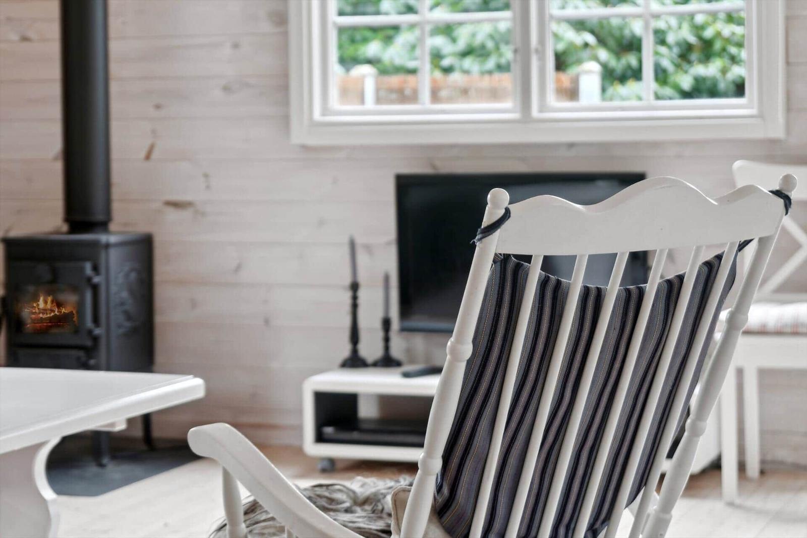 Ein weißer Sessel mit Streifenkissen steht vor einem Fenster. Im Hintergrund ist ein Kaminofen mit brennendem Feuer.