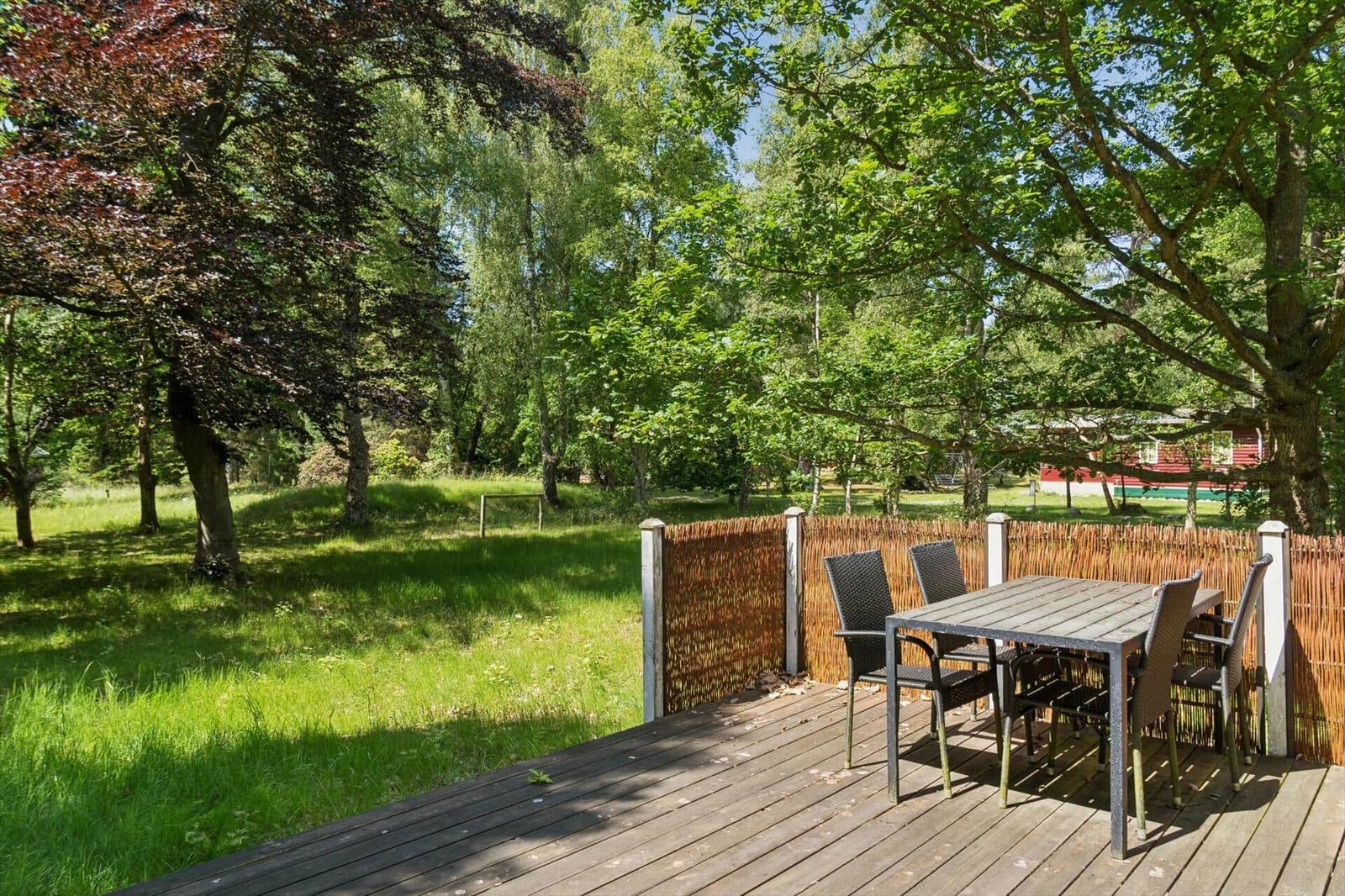 Terrasse mit Tisch und Stühlen unter Bäumen. Grasfläche und Zaun im Hintergrund.