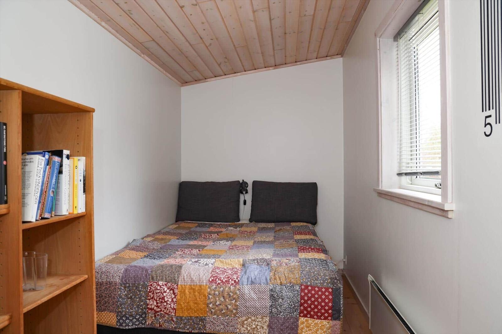 Schlafzimmer mit Bett, Bücherregal und Fenster. Holzdecke und gemustertes Bettlaken.