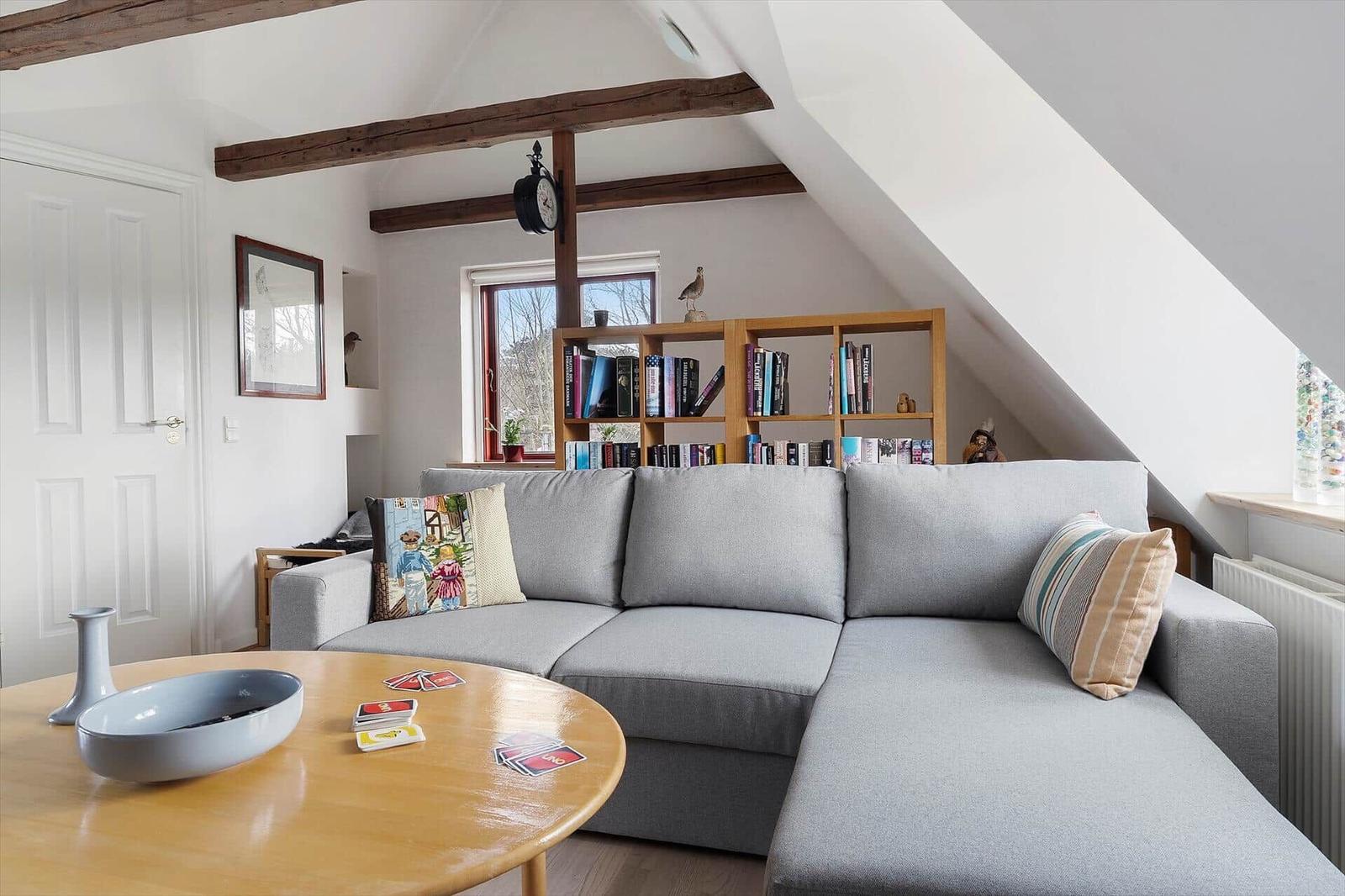 Wohnzimmer mit grauem Sofa, Holztisch und Bücherregal unter Dachschrägen.