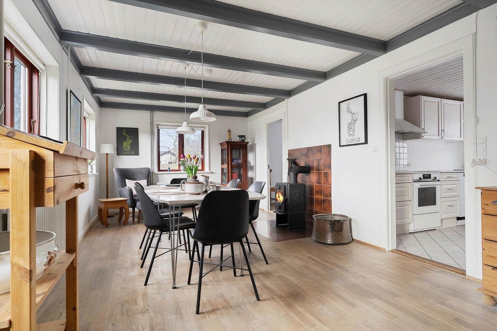 Esszimmer mit Holzboden, Esstisch und Kaminofen. Küche ist angrenzend.