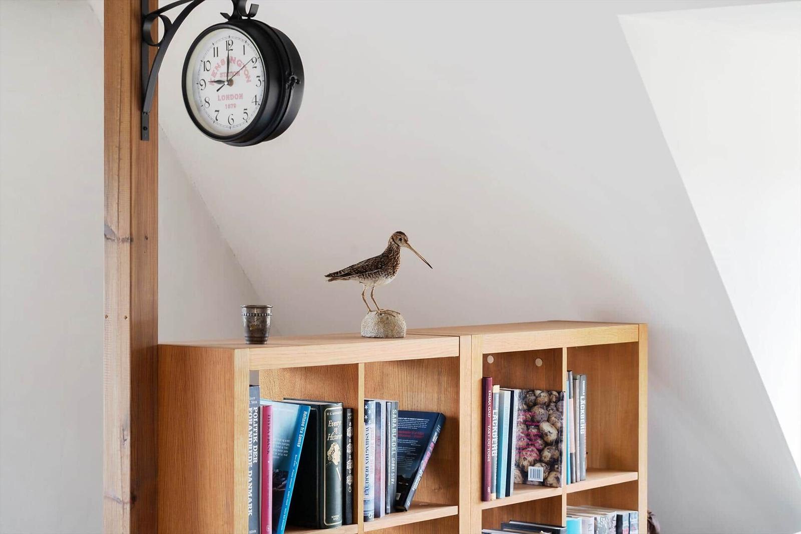 Holzregal mit Büchern, Vogeldekoration und Wanduhr an schräger Wand.