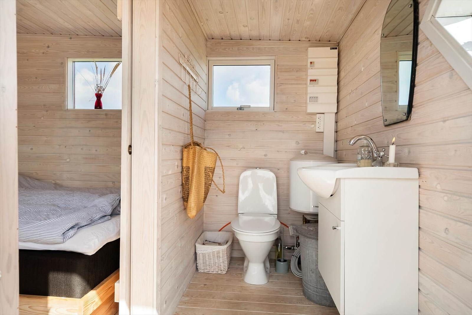 Badezimmer mit Toilette, Waschbecken und Fenster. Holzwände und -boden. Bett im Hintergrund.