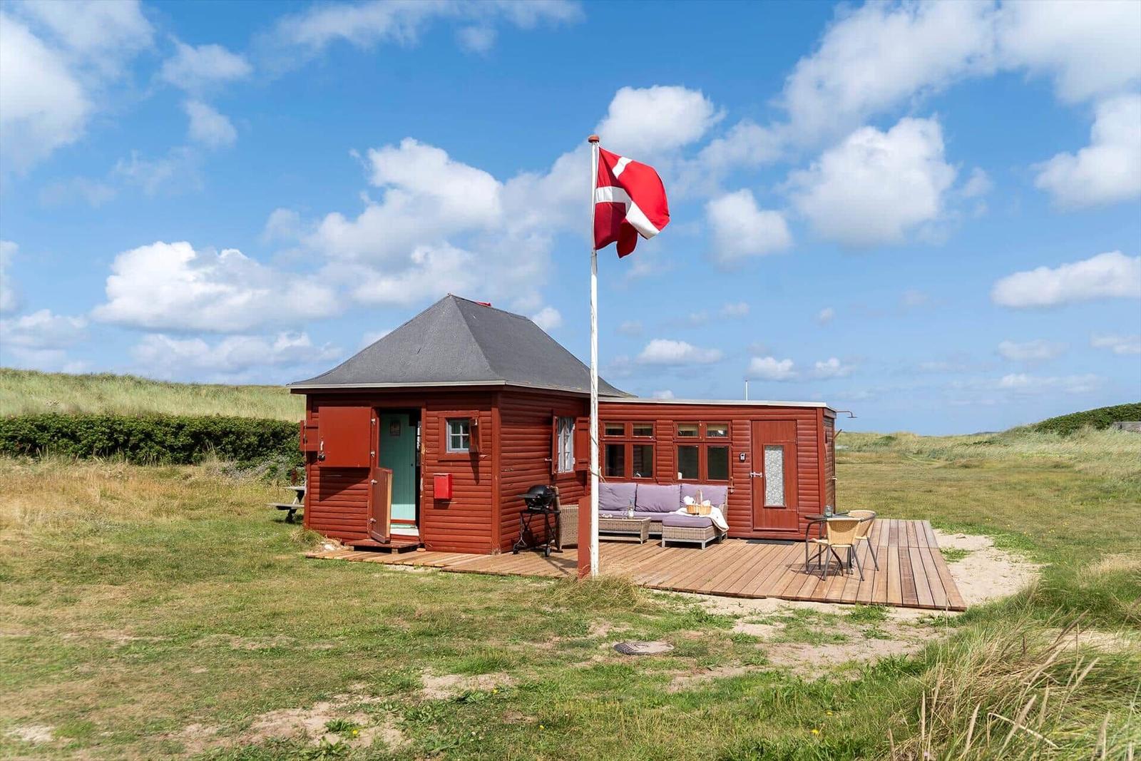Rotes Holzhaus mit Terrasse, Grill und dänischer Flagge auf grünem Grund.
