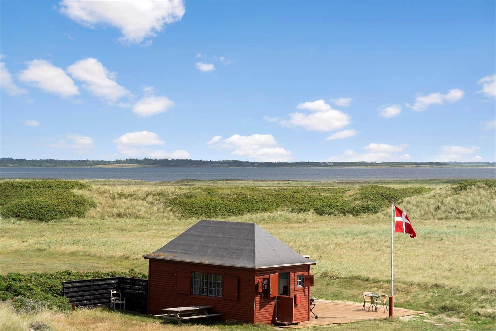 Rotes Holzhaus mit Terrasse, Blick auf Wasser und Dünen. Daneben steht eine Flagge.