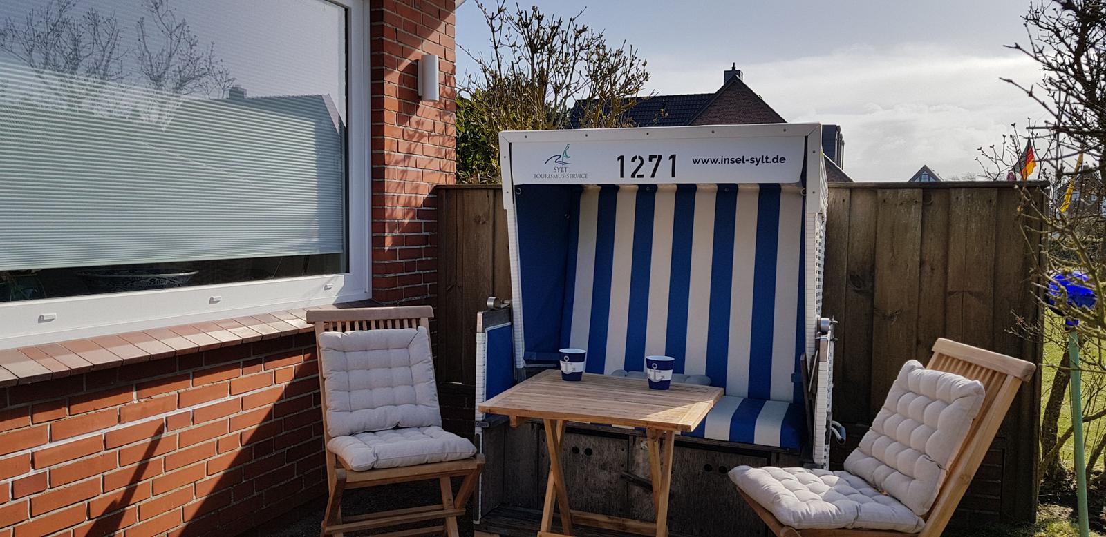 Terrasse mit Strandkorb, Tisch und zwei Stühlen vor Hauswand.