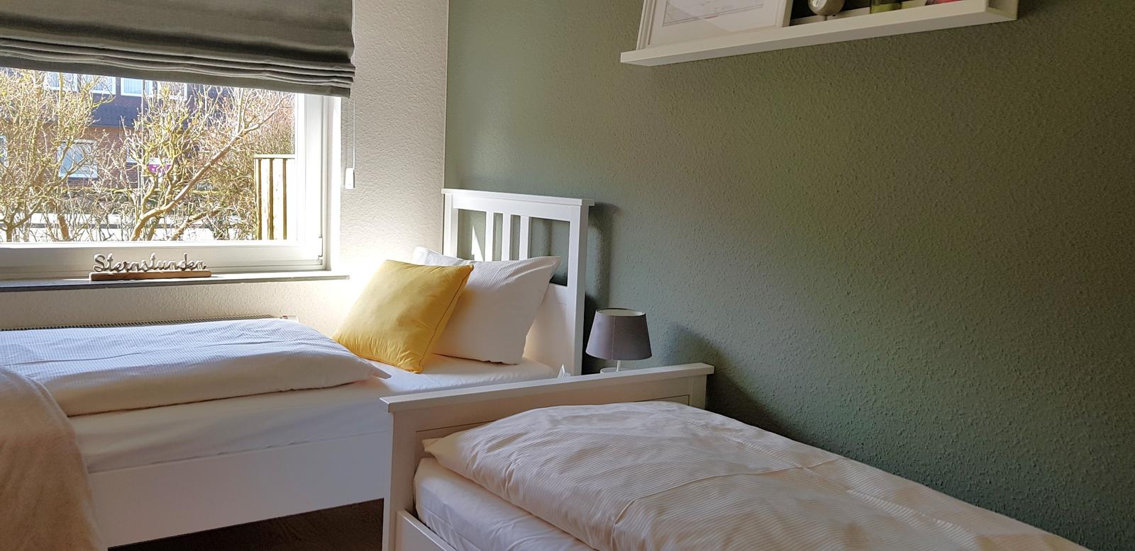 Zwei Betten mit weißen Bettwäschen und einem gelben Kissen. Grüne Wand und Fenster mit Blick nach draußen.