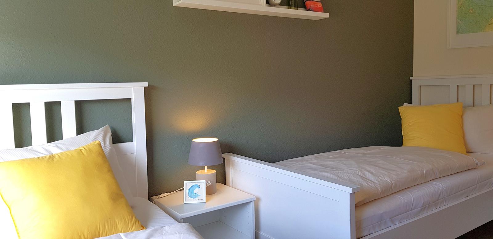 Zwei Betten mit weißen Rahmen und gelben Kissen. Zwischen ihnen steht ein Nachttisch mit Lampe.