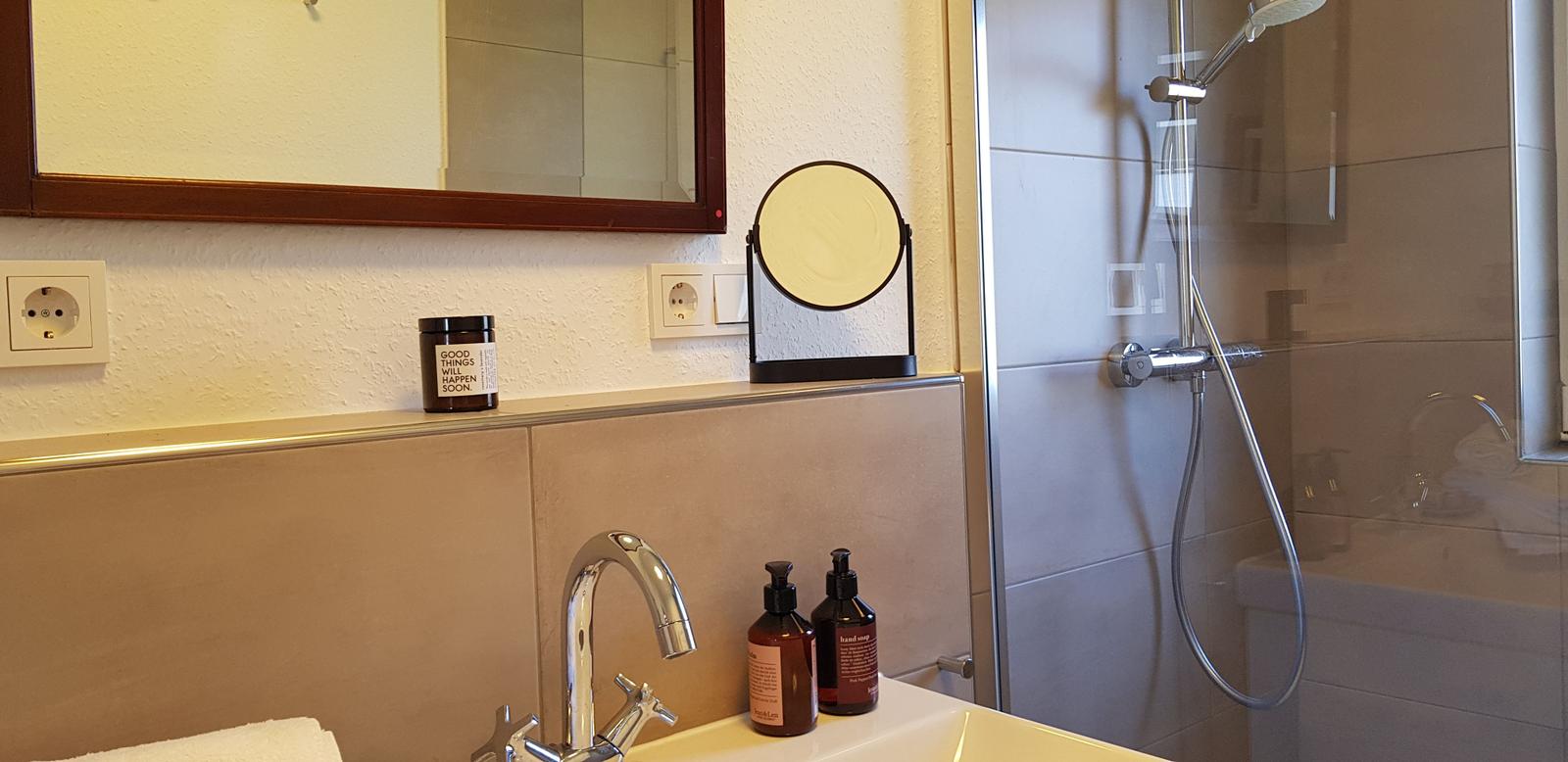 Bad mit Dusche, Waschbecken und Spiegel. Auf der Ablage stehen Seifen und ein Kerzenhalter.