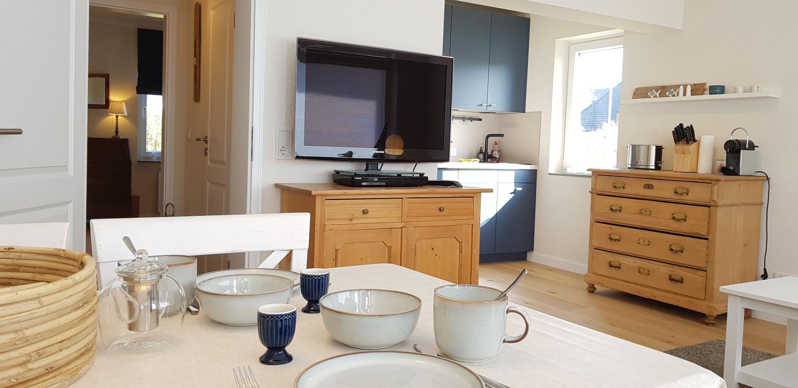 Esstisch mit Geschirr, Fernseher und Küchenbereich im Hintergrund.