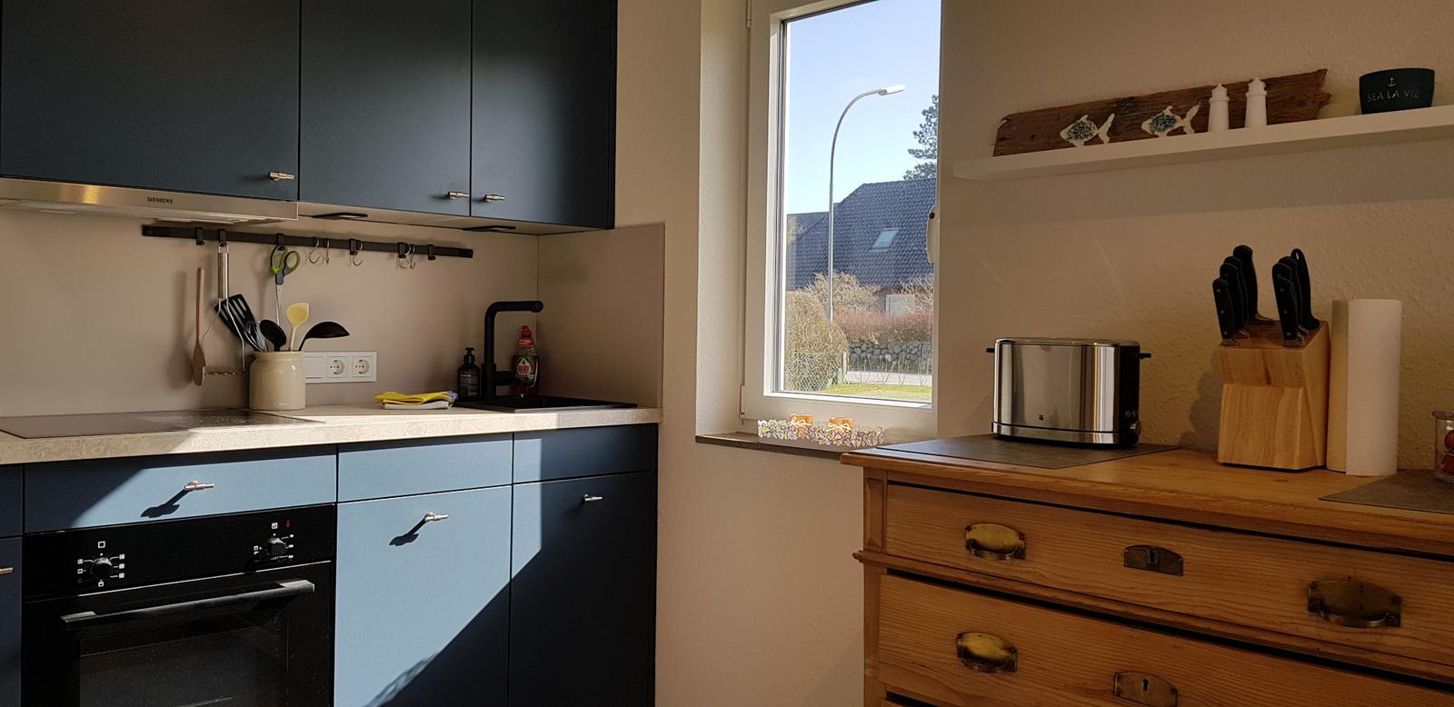 Küche mit dunklen Schränken, Fenster und Holzschrank. Auf dem Fensterbrett liegen Süßigkeiten.