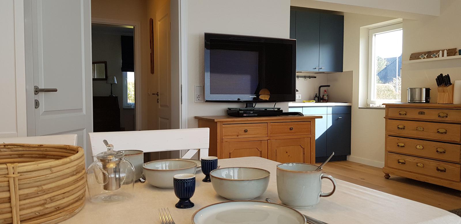 Esszimmer mit Tisch, Schüsseln und Kaffeetasse. Küche und TV im Hintergrund.