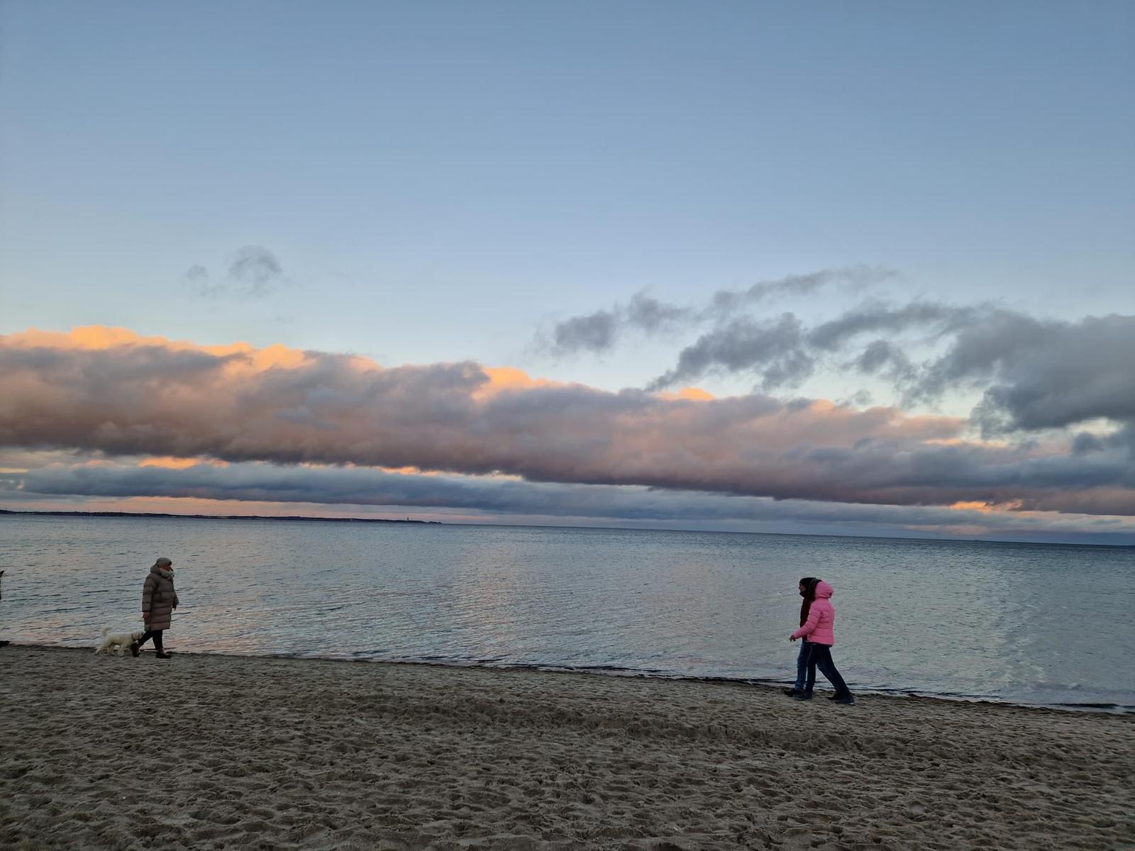 Zwei Personen und ein Hund spazieren am Strand entlang bei Sonnenuntergang.