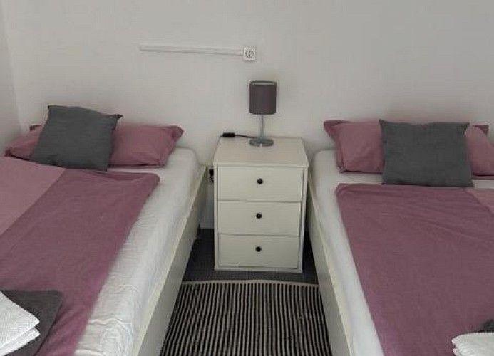 Zwei Einzelbetten mit rosa und grauen Kissen und einem weißen Nachttisch dazwischen.